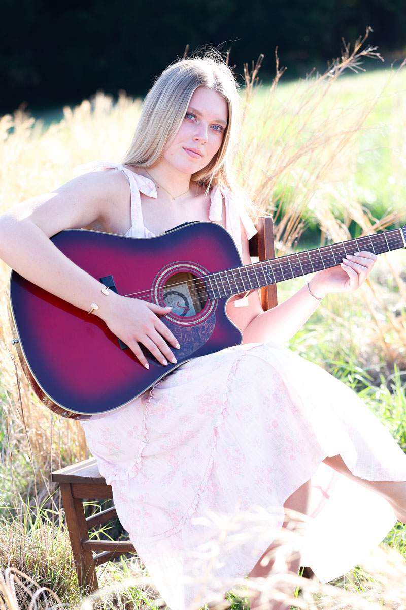 Senior portrait of a young woman playing an acoustic guitar in a golden Texas field at sunset