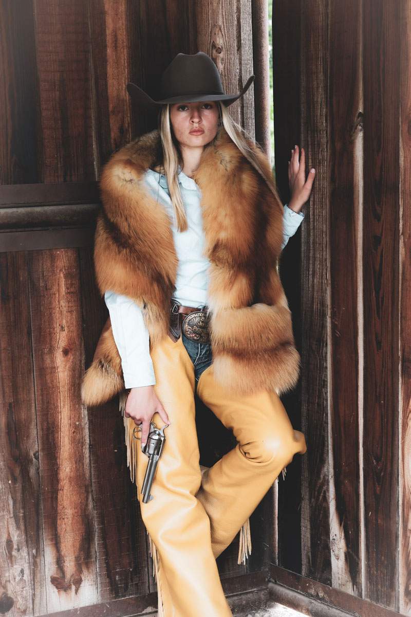 Editorial Western portrait of a woman in a cowboy hat, fur stole and leather chaps in a weathered barn doorway